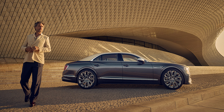 Bentley Gold Coast (Australia) Bentley Flying Spur Mulliner sedan in Rose Gold over Magnetic duotone paint parked by a curved wall with person in suit jacket standing on the left